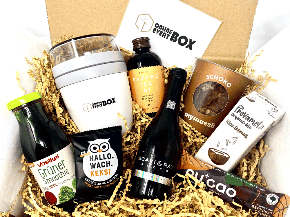 Pictured is an online event box filled with a reusable granola yogurt cup, crunchy chocolate granola, healthy green smoothie, invigorating cold brew coffee, refreshing Rice Coconut drink, sparkling prosecco, delicious espresso crunch chocolate bar, greeting card and a caffeinated Hallo.Wach.Keks!