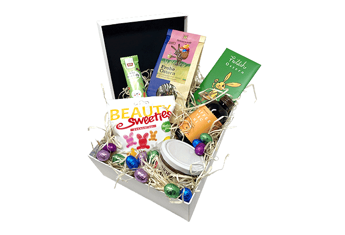 Eine Online Event Box Osterbox gefüllt mit Heu, Schokoladeneiern, zuckerfreien Fruchtgummi-Häschen, saftigem Kuchen und Schokolade in verschiedenen osterlichen Formen.
